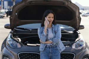 Žena, která telefonuje a je opřená zepředu o auto se zvednutou kapotou.