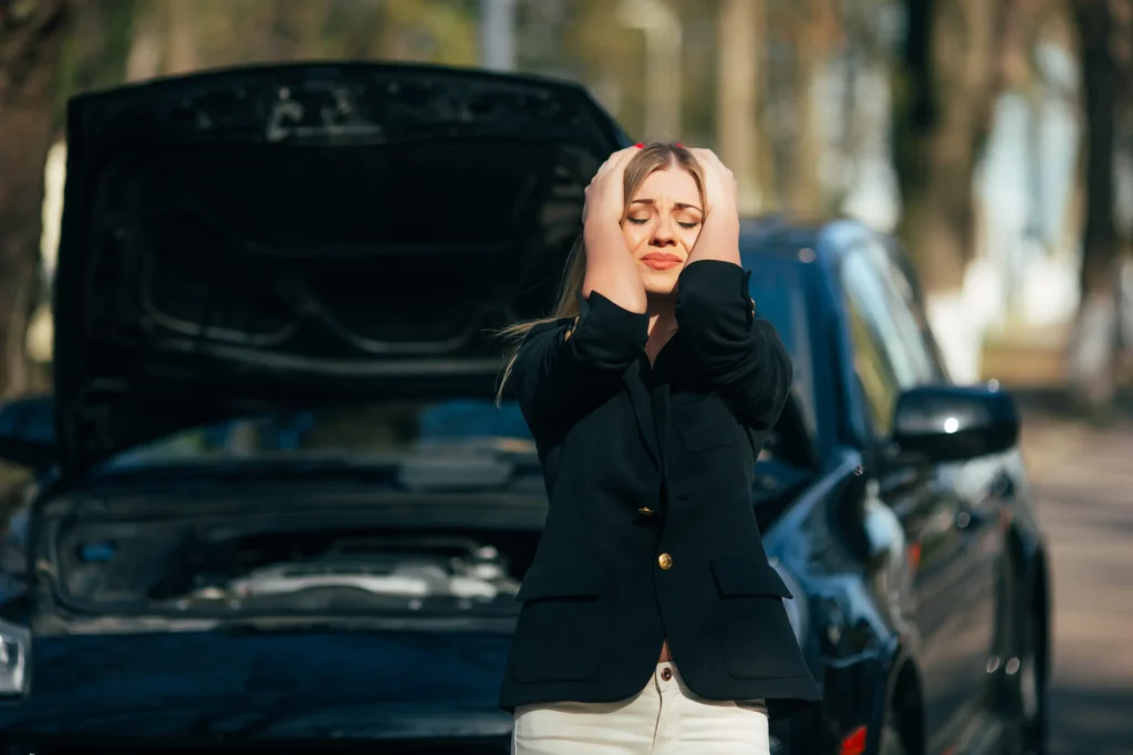 Frustrovaná mladá žena stojí před autem s otevřenou kapotou, drží si hlavu v dlaních a vypadá zoufale, auto má zjevně poruchu.