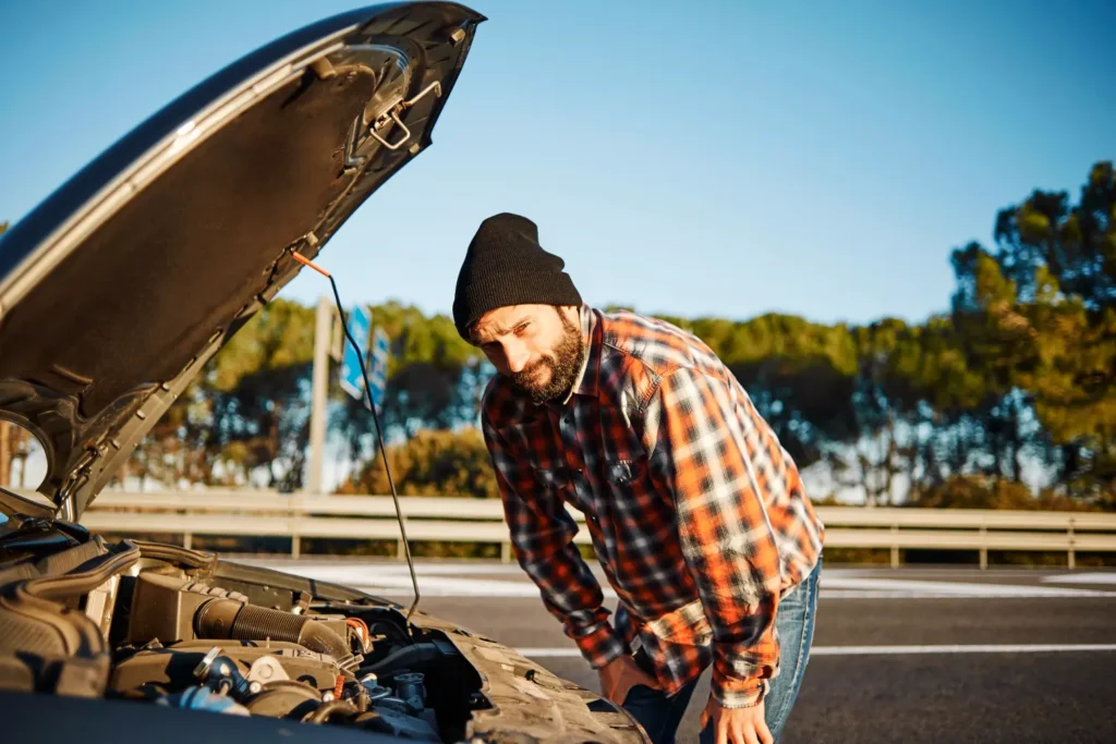 Vousatý muž v kostkované košili a čepici kontroluje motor auta s otevřenou kapotou na kraji silnice.